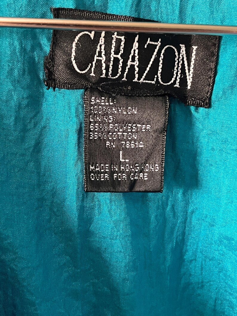 Pode incluir: Uma jaqueta azul turquesa com uma etiqueta preta que diz "CABAZON" e lista o conte&uacute;do do tecido como 100% nylon, 65% poli&eacute;ster e 35% algod&atilde;o. A etiqueta tamb&eacute;m diz "Feito em Hong Kong" e "Virar para o cuidado".
