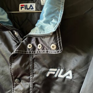 Op de afbeelding: Zwarte Fila windbreaker jas met een lichtblauwe voering en witte stiksels. De jas heeft een ritssluiting en een drukknoop aan de onderkant. Het Fila-logo is in wit geborduurd op de voorkant van de jas.