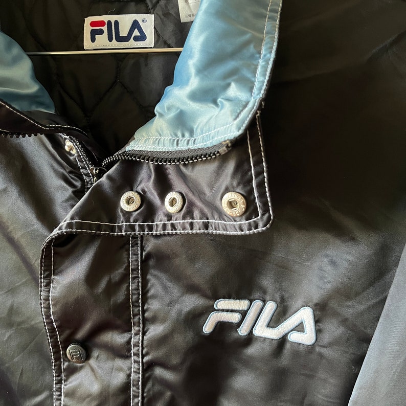 Op de afbeelding: Zwarte Fila windbreaker jas met een lichtblauwe voering en witte stiksels. De jas heeft een ritssluiting en een drukknoop aan de onderkant. Het Fila-logo is in wit geborduurd op de voorkant van de jas.