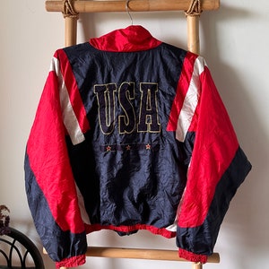 Könnte beinhalten: Eine blaue, rote und weiße Windbreaker-Jacke mit dem Wort "USA" auf der Rückseite gestickt. Die Jacke hat einen Reißverschluss und einen Stehkragen.