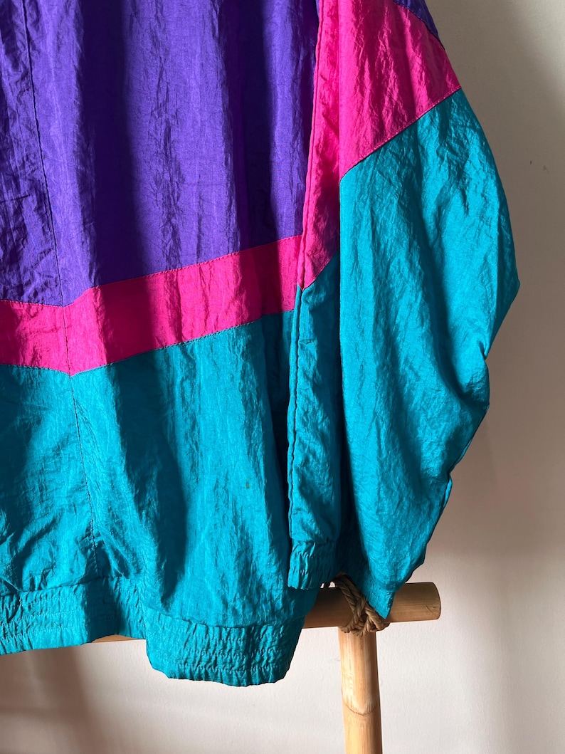 Pode incluir: Uma jaqueta corta-vento vintage com design de blocos de cores. A jaqueta &eacute; feita de tecido de nylon leve e apresenta um esquema de cores roxo, rosa e turquesa. A jaqueta tem fecho de correr e gola alta.