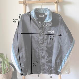 Op de afbeelding: Zwarte Fila windbreaker-jas met witte en blauwe accenten. De jas heeft een drukknoopsluiting en een trekkoord aan de onderkant. Hij is 26 inch breed en 30 inch lang.