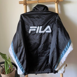 Op de afbeelding: Zwarte Fila windbreaker-jas met witte en lichtblauwe accenten. De jas heeft een ritssluiting en een groot Fila-logo op de rug.