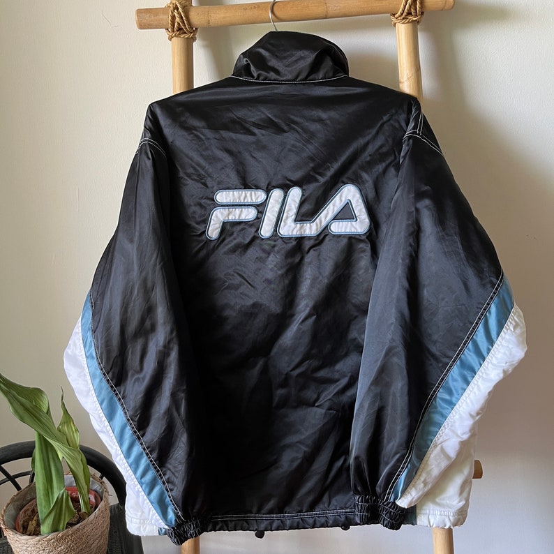 Op de afbeelding: Zwarte Fila windbreaker-jas met witte en lichtblauwe accenten. De jas heeft een ritssluiting en een groot Fila-logo op de rug.