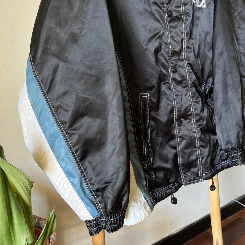 Op de afbeelding: Zwarte satijnen jas met witte en blauwe strepen op de mouwen. De jas heeft een drukknoopsluiting en een wit stikdetail.