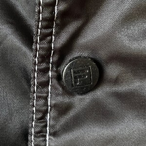 Op de afbeelding: Close-up van een zwarte knoop met een witte gestikte naad op een zwarte stoffen achtergrond. De knoop heeft een verhoogd vierkant met de letter "F" erin.