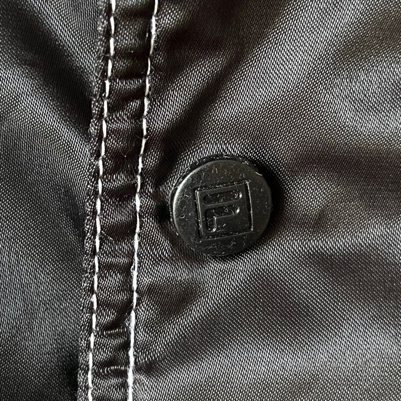 Op de afbeelding: Close-up van een zwarte knoop met een witte gestikte naad op een zwarte stoffen achtergrond. De knoop heeft een verhoogd vierkant met de letter "F" erin.