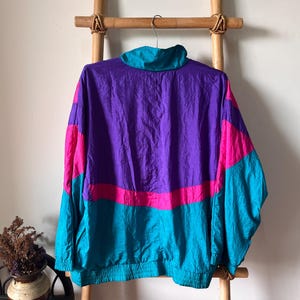 Pode incluir: Uma jaqueta corta-vento de cor roxa, rosa e turquesa com fecho de correr. A jaqueta tem um colar de p&eacute; e mangas compridas.