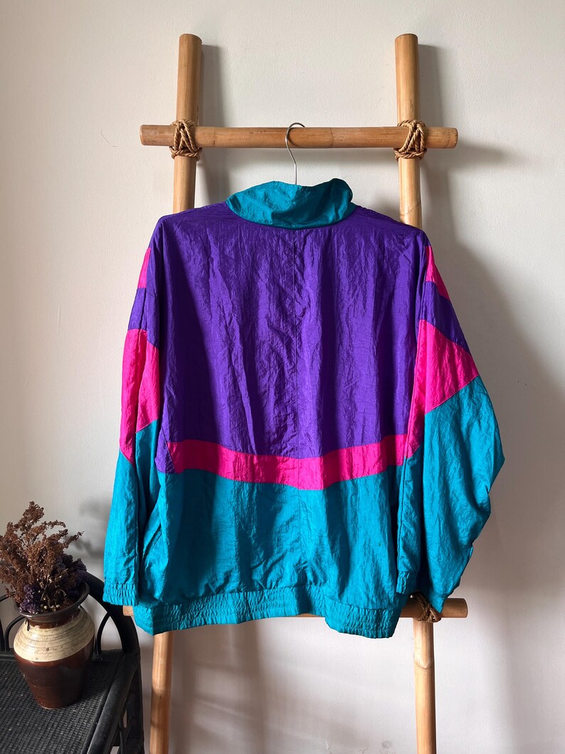 Pode incluir: Uma jaqueta corta-vento de cor roxa, rosa e turquesa com fecho de correr. A jaqueta tem um colar de p&eacute; e mangas compridas.