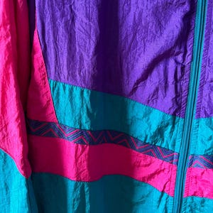 Pode incluir: Uma jaqueta colorida com fecho de correr. A jaqueta &eacute; feita de tecido de nylon e tem um padr&atilde;o geom&eacute;trico em tons de rosa, roxo e turquesa.