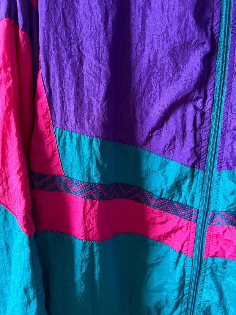 Pode incluir: Uma jaqueta colorida com fecho de correr. A jaqueta &eacute; feita de tecido de nylon e tem um padr&atilde;o geom&eacute;trico em tons de rosa, roxo e turquesa.