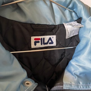 Op de afbeelding: Een blauw en zwart Fila jack met een gewatteerde voering. Het jack heeft een ritssluiting en een drukknoop aan de kraag. Het Fila logo is zichtbaar aan de binnenkant van het jack.