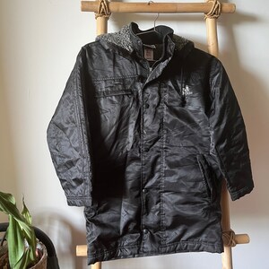 May include: Black hooded jacket with a fleece lining and a logo on the left chest. The jacket has a zipper closure and multiple pockets.