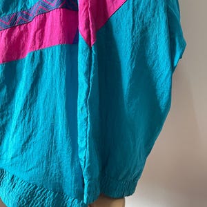 Pode incluir: Uma jaqueta corta-vento azul turquesa, rosa e roxa com um padr&atilde;o geom&eacute;trico. A jaqueta tem um fecho de correr e um capuz com cord&atilde;o.