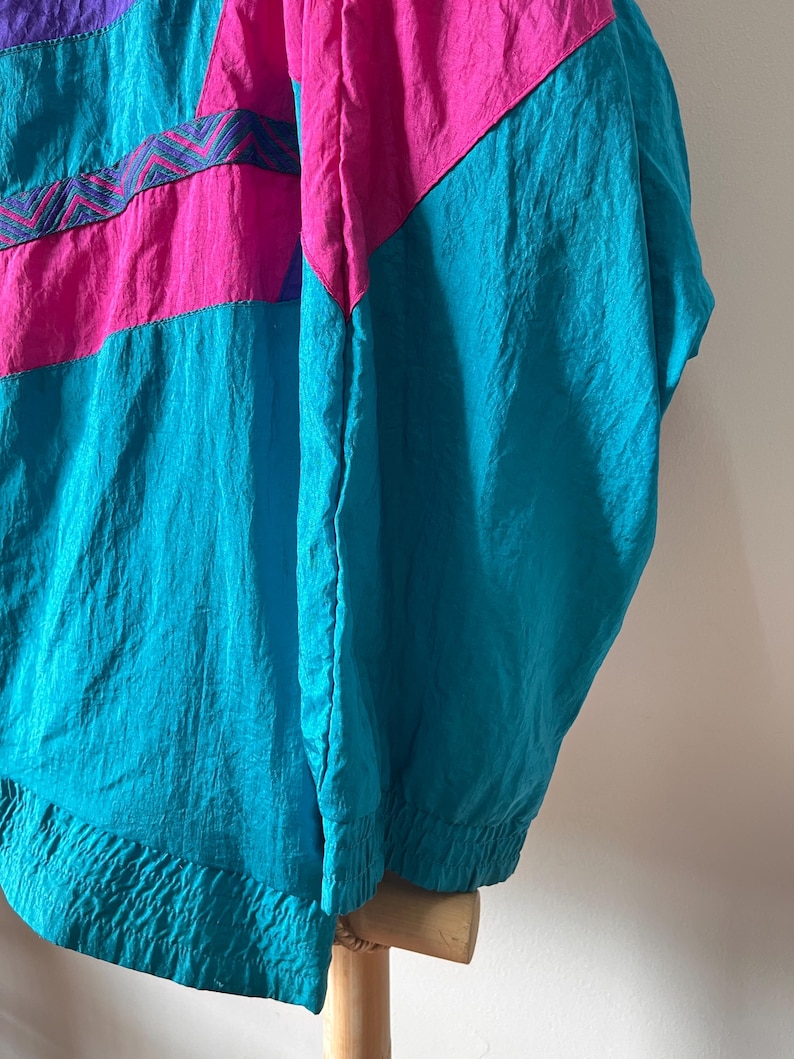 Pode incluir: Uma jaqueta corta-vento azul turquesa, rosa e roxa com um padr&atilde;o geom&eacute;trico. A jaqueta tem um fecho de correr e um capuz com cord&atilde;o.