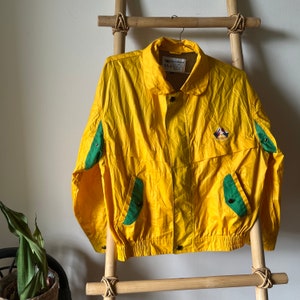 Puede incluir: Una chaqueta de nylon amarilla con detalles verdes y un logotipo en el pecho. La chaqueta tiene cierre de cremallera y cintura con cordón.