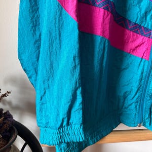 Pode incluir: Uma jaqueta corta-vento azul turquesa, rosa e roxa com fecho de correr. A jaqueta tem um padr&atilde;o geom&eacute;trico com um design em chevron.
