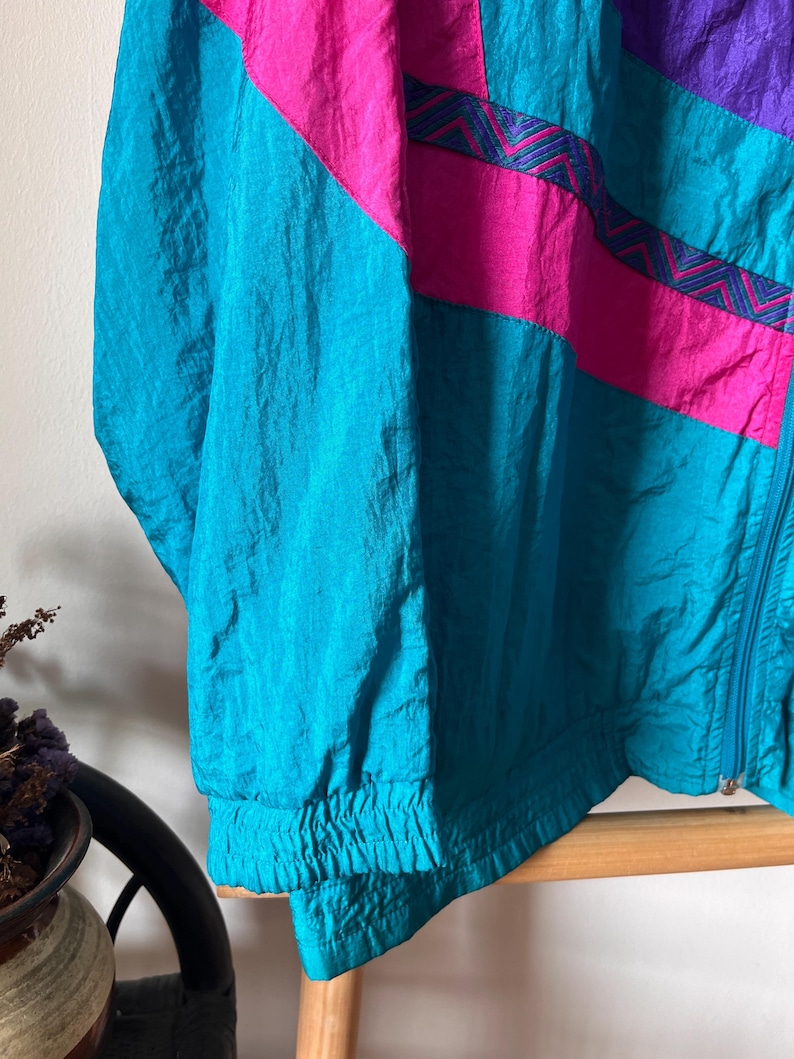 Pode incluir: Uma jaqueta corta-vento azul turquesa, rosa e roxa com fecho de correr. A jaqueta tem um padr&atilde;o geom&eacute;trico com um design em chevron.