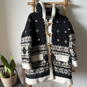 May include: Black and white patterned hooded jacket with a white sherpa lining and wooden toggle closures. The jacket has two front pockets.