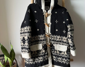 May include: Black and white patterned hooded jacket with a white sherpa lining and wooden toggle closures. The jacket has two front pockets.