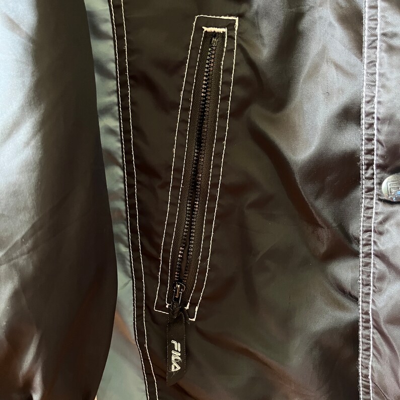Op de afbeelding: Close-up van een zwart jack met een witte rits en witte stiksels. De rits heeft een Fila-logo onderaan.