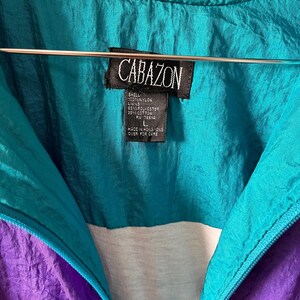 Pode incluir: Uma jaqueta corta-vento turquesa e roxa com um z&iacute;per branco e uma etiqueta preta que diz "CABAZON" com as seguintes informa&ccedil;&otilde;es: "SHELL: NYLON LINING: 65% POLYESTER 35% COTTON RV 78610 L MADE IN HONG KONG OVER FOR CARE"