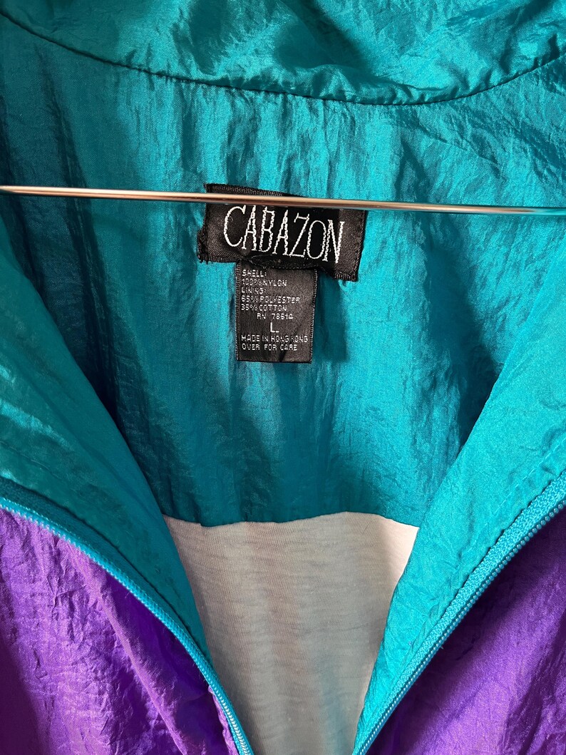 Pode incluir: Uma jaqueta corta-vento turquesa e roxa com um z&iacute;per branco e uma etiqueta preta que diz "CABAZON" com as seguintes informa&ccedil;&otilde;es: "SHELL: NYLON LINING: 65% POLYESTER 35% COTTON RV 78610 L MADE IN HONG KONG OVER FOR CARE"