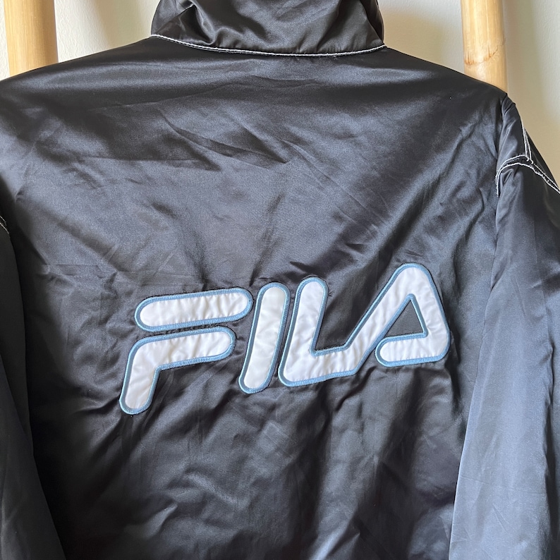Op de afbeelding: Zwarte satijnen jas met een geborduurd FILA-logo in wit en lichtblauw op de rug.