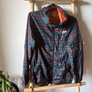 Puede incluir: Una chaqueta de color azul, verde y rojo con estampado de paisley, con una cremallera negra y capucha. La chaqueta tiene una pequeña etiqueta naranja con el texto "TITTIKA" en ella.