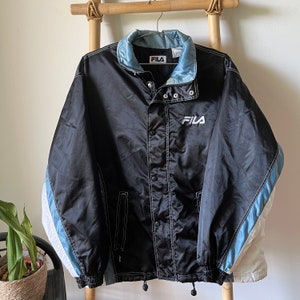 Op de afbeelding: Zwarte Fila windbreaker-jas met witte en blauwe accenten. De jas heeft een drukknopsluiting en een capuchon met trekkoord.