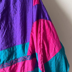 Pode incluir: Uma jaqueta colorida com um esquema de cores roxo, rosa e turquesa. A jaqueta tem um design geom&eacute;trico com um padr&atilde;o em ziguezague na se&ccedil;&atilde;o turquesa.