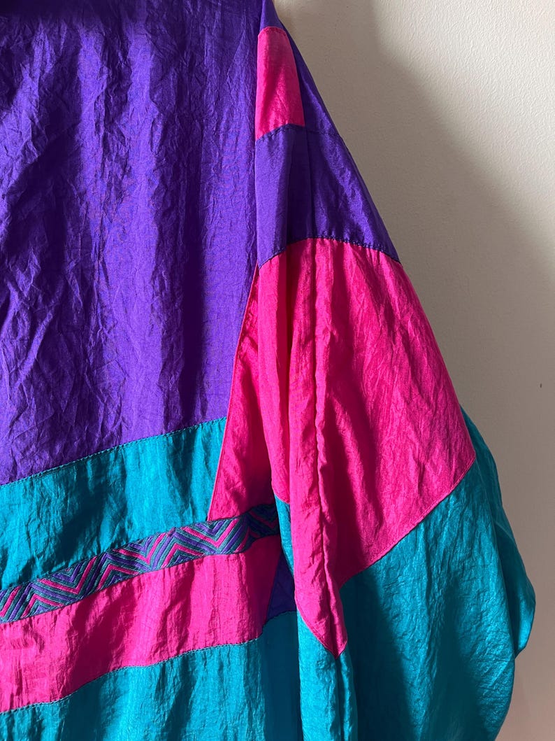 Pode incluir: Uma jaqueta colorida com um esquema de cores roxo, rosa e turquesa. A jaqueta tem um design geom&eacute;trico com um padr&atilde;o em ziguezague na se&ccedil;&atilde;o turquesa.
