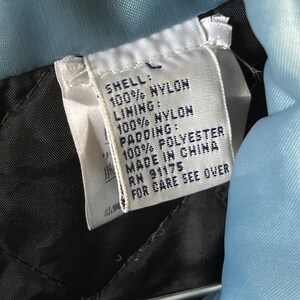 Op de afbeelding: Een wit label met zwarte tekst die de stofsamenstelling van een jas beschrijft. Het label luidt "Shell: 100% Nylon, Lining: 100% Nylon, Padding: 100% Polyester, Made in China, RN 91175, For Care See Over."