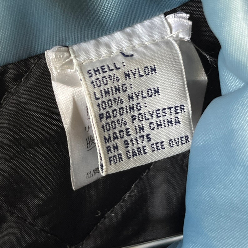Op de afbeelding: Een wit label met zwarte tekst die de stofsamenstelling van een jas beschrijft. Het label luidt "Shell: 100% Nylon, Lining: 100% Nylon, Padding: 100% Polyester, Made in China, RN 91175, For Care See Over."