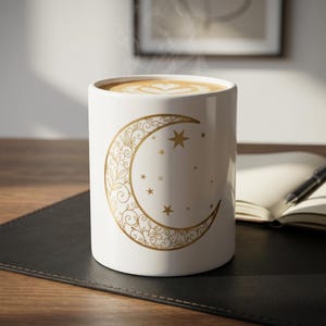 May include: A white ceramic mug featuring a gold-colored crescent moon design filled with intricate floral filigree patterns and small stars. The mug is filled with a hot beverage topped with latte art, emitting a faint wisp of steam from the top.