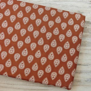 Op de afbeelding: Een gevouwen stuk stof in een diepe terracotta oranje kleur met een herhalend patroon van witte druppelvormige motieven. Elke druppel bevat een klein, centraal bloemmotief met vijf bloemblaadjes, gerangschikt in een consistent raster.