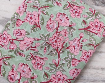 Op de afbeelding: Gevouwen stof met een bloemenpatroon. De stof heeft een lichtgroene achtergrond met een herhalend patroon van roze bloemen en rode stelen. De bladeren zijn groen.