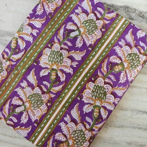 Könnte beinhalten: Gefalteter violetter Stoff mit floralem Muster. Das Design zeigt große weiße und gelbe Blumen mit grünen Akzenten auf einem violetten Hintergrund. Grüne und weiße Streifen umranden die floralen Motive.