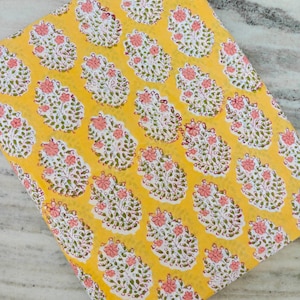 May include: A folded piece of yellow fabric with a repeating floral pattern. The pattern features pink flowers and green leaves on a white background. The fabric is likely cotton or a similar material.