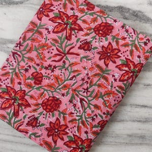 Peut inclure: Un tissu plié avec un fond rose et un motif floral. Le motif présente de grandes fleurs rouges, de plus petits boutons rouges, des feuilles vertes et des accents orange. Le tissu est probablement en coton ou similaire.