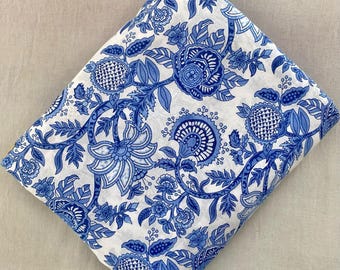 May include: Folded white fabric with a blue floral pattern. The design features large, stylized flowers, leaves, and vines in varying shades of blue. The fabric is folded diagonally, showcasing the repeating pattern.