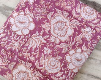 May include: Folded fabric with a floral pattern. The fabric features large, stylized flowers in white and light brown against a mauve background. The design includes leaves and smaller floral elements, creating a detailed and textured appearance.