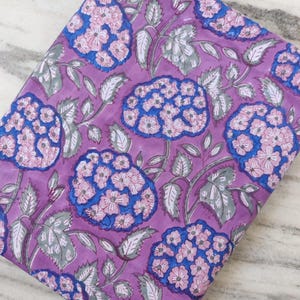 Peut inclure: Tissu plié avec un motif floral. Le motif présente de grandes fleurs stylisées dans des tons de bleu et de rose sur un fond violet. Les feuilles sont d'un gris-vert sourd. Le tissu est probablement en coton ou dans une matière similaire.