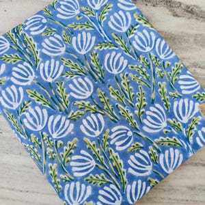 May include: A folded piece of fabric with a repeating floral pattern. The design features white flowers with green stems and leaves on a light blue background. The fabric appears to be cotton or a similar material, suitable for various sewing projects.