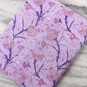 Op de afbeelding: Gevouwen stof met een herhalend bloemenpatroon. Het ontwerp toont gestileerde granaatappels en bladeren in tinten paars en roze tegen een lichtpaarse achtergrond. De stof is waarschijnlijk katoen of een vergelijkbaar materiaal, geschikt voor naaiprojecten.