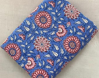 May include: Folded blue fabric with a floral pattern. The design features large pink and red flowers, smaller pink blossoms, and white leaves on a blue background. The fabric is likely cotton or a similar material.