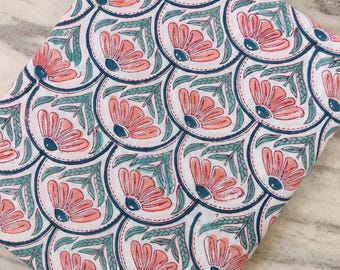 May include: Folded white fabric with a repeating pattern of coral-colored flowers, teal leaves, and navy blue outlines. The floral design is arranged in a scale-like pattern. The fabric is likely cotton or linen.