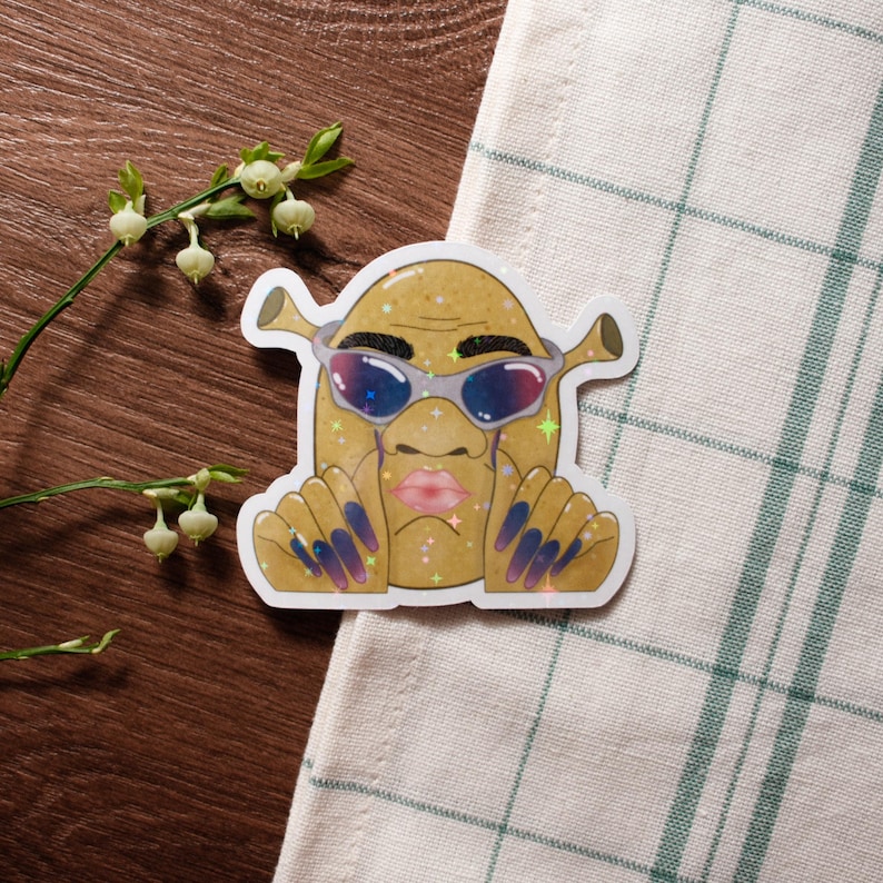 May include: A sticker featuring a cartoon character with a tan complexion, wearing sunglasses and long, blue fingernails. The character has pink lips and is surrounded by glitter. The sticker is on a wooden surface.