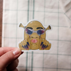 May include: A holographic sticker featuring a cartoon character with a potato-like body, Shrek-like ears, and sunglasses. The character has a pink lip, tears, and long, dark blue and purple nails. The sticker is held by a hand.