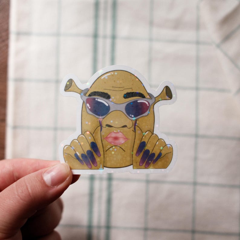 May include: A holographic sticker featuring a cartoon character with a potato-like body, Shrek-like ears, and sunglasses. The character has a pink lip, tears, and long, dark blue and purple nails. The sticker is held by a hand.